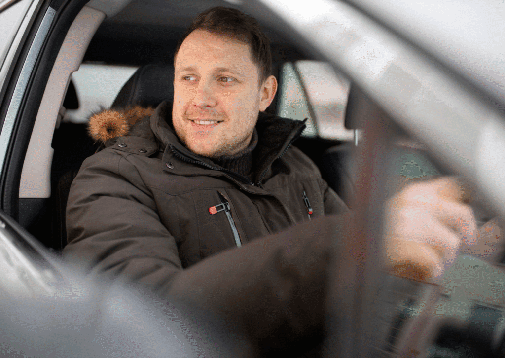 Smiling man in a winter coat driving a car, viewed through the open window.